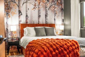Een slaapkamer met een bed, kussens, een nachtkastje met een lamp, een raam met gordijnen, en een wand met behangpapier met een natuurmotief.