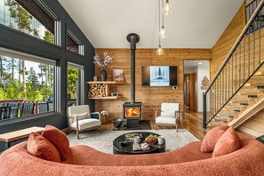 A modern living room with a fireplace, wooden walls, and a large window overlooking trees. - Family retreat + Game Room, Fire Pit & Views | Near RMNP+ Grand Lake (Grand Lake)