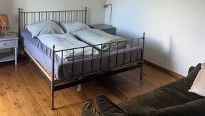 Un lit simple avec un cadre en métal, une table de chevet, une lampe et un canapé de couleur sombre, dans une pièce au plancher en bois et aux murs blancs.