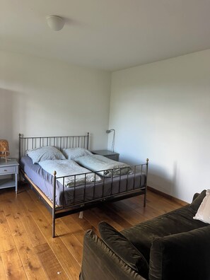 A single bed with a metal frame, a bedside table, a lamp, and a dark-colored sofa in a room with wooden flooring and white walls. - Cozy Vacation Rental at a Horse Farm (Poggensee)