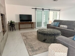 Un salon moderne dotĂ© dâun tĂ©lĂ©viseur Ă Ă©cran plat, dâun canapĂ©, de poufs, dâun tapis et dâune porte-fenĂȘtre coulissante.