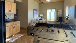 A room with wooden ceiling, a foosball table, a kitchen with wooden cabinets, and a seating area with a sofa and a TV.