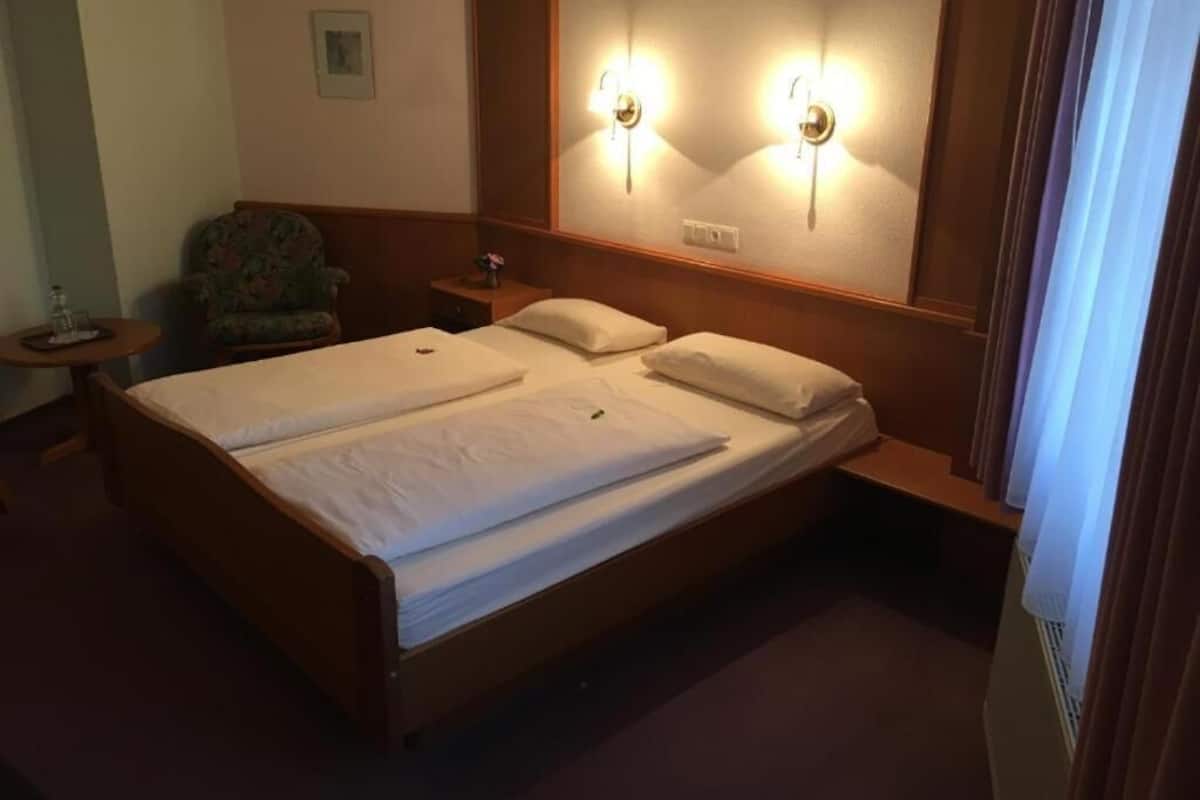 A double bed with two pillows, a small round table with a bottle, a chair, and wall-mounted light fixtures.