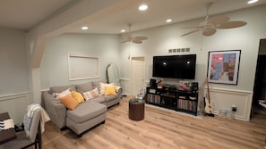 A living room with a grey sofa, ceiling fans, a TV, a bookshelf, and a guitar. - All Sports Lakefront Sunset w/Deck, Firepit, Dock (Ortonville)