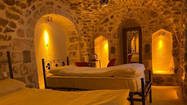 Una habitación con paredes de piedra, dos camas, un espejo y luces fijadas a la pared.