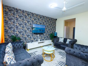 A living room with a stone-patterned wall, a flat-screen TV, a ceiling fan, and a sofa set. - Shanzu Serenity:  Your 3-Bedroom Coastal Haven Near PrideInn Paradise .#Msa.. (Mombasa)