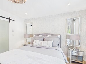 A modern bedroom with a large bed, bedside lamps, a glass side table, and a mirror. - Luxury 3BR Townhome with Garden Oasis (Victoria)