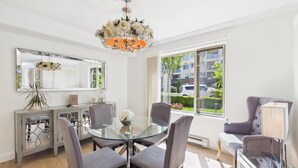 A modern dining area with a glass table, four grey chairs, a chandelier, a large window with a view of a residential area, and a mirrored cabinet. - Luxury 3BR Townhome with Garden Oasis (Victoria)