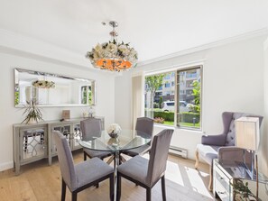 A modern dining area with a glass table, four grey chairs, a chandelier, a large window with a view of a residential area, and a mirrored cabinet. - Luxury 3BR Townhome with Garden Oasis (Victoria)
