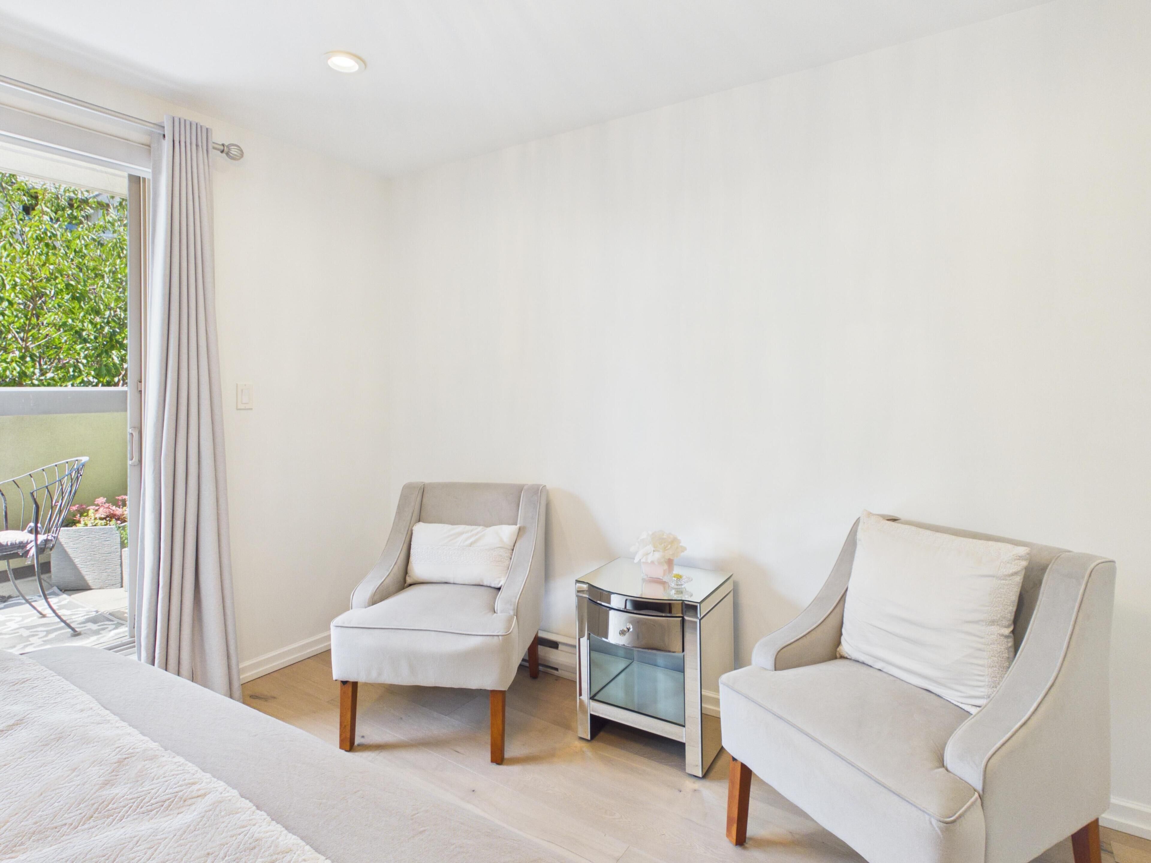 Ein Zimmer mit zwei Sesseln, einem kleinen Nachttisch und einem großen Fenster mit Vorhängen.