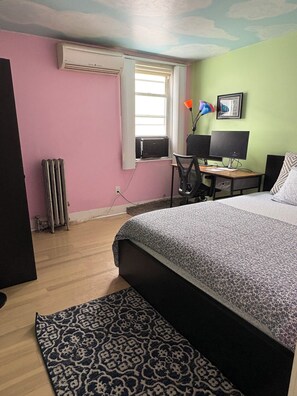 A bedroom with a bed, a desk with a computer, a radiator, and a window.