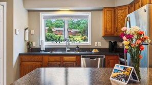 A kitchen with wooden cabinets, a stainless steel refrigerator, a dishwasher, and a window with a view of houses. - Fox Lair by Avantstay Oversized Jetted Tub + Grilling Deck Heated Pool + Recreation Center (North Creek)