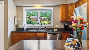 A kitchen with wooden cabinets, a stainless steel refrigerator, a dishwasher, and a window with a view of houses.