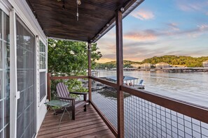 Une terrasse en bois avec une chaise et une table, donnant sur une rivière où l’on aperçoit des bateaux et des bâtiments au loin.