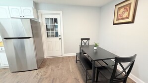 Eine Küche mit Kühlschrank, einer weißen Tür, einem Esstisch mit Stühlen und einem an der Wand hängenden Bild.