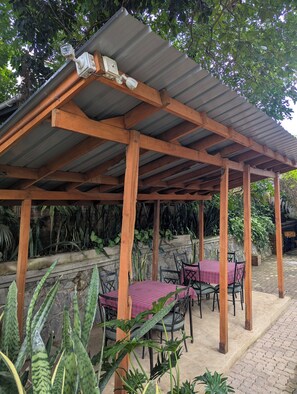 A covered outdoor seating area with tables and chairs, surrounded by plants and a stone wall. - Bamboo Savanna Villas (Arusha)