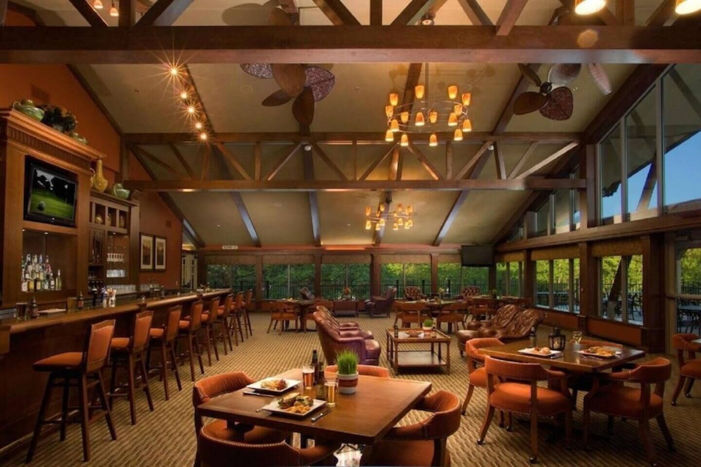 Amplio espacio interior con techos altos, vigas de madera y grandes ventanales que ofrecen vistas al exterior. Varias zonas de estar con mesas y sillas, además de una zona de bar con mostrador y taburetes.