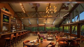 Amplio espacio interior con techos altos, vigas de madera y grandes ventanales que ofrecen vistas al exterior. Varias zonas de estar con mesas y sillas, además de una zona de bar con mostrador y taburetes.