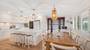 Una cuina moderna amb armaris blancs, una illa central amb tamborets, il·luminació penjant i una zona de menjador amb taula i cadires de fusta.