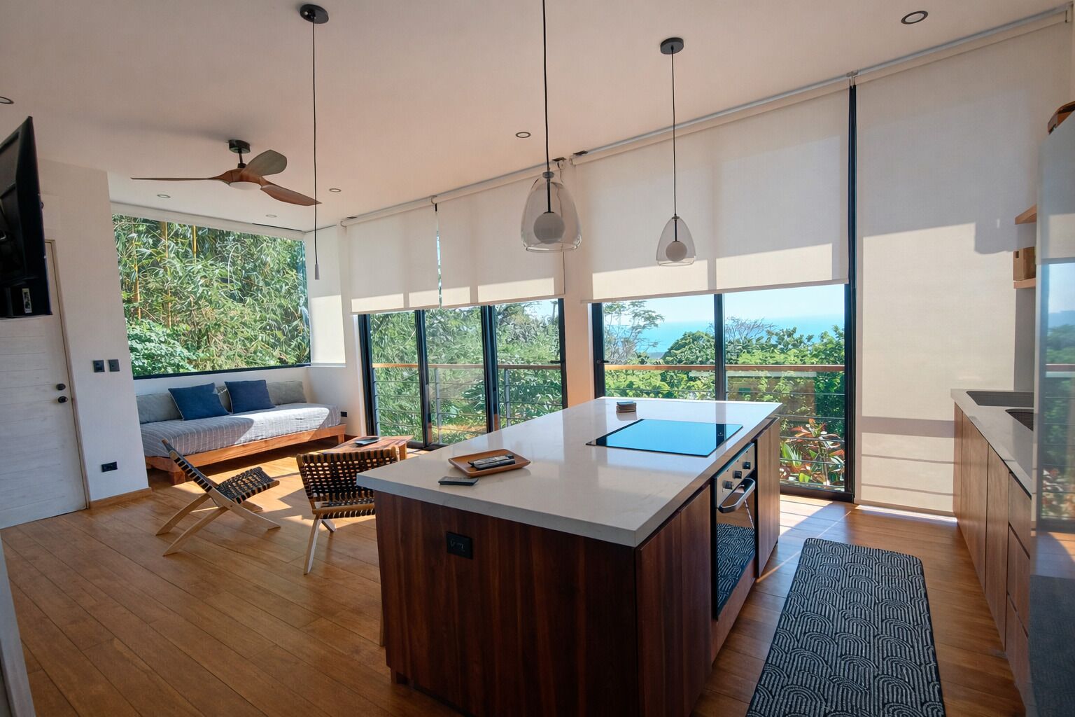 A modern kitchen with a central island, large windows offering a view of greenery, and a seating area with a sofa and chairs.