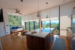 Eine moderne Küche mit einer zentralen Kochinsel, großen Fenstern mit Blick auf Grünflächen und einer Sitzecke mit Sofa und Sesseln.