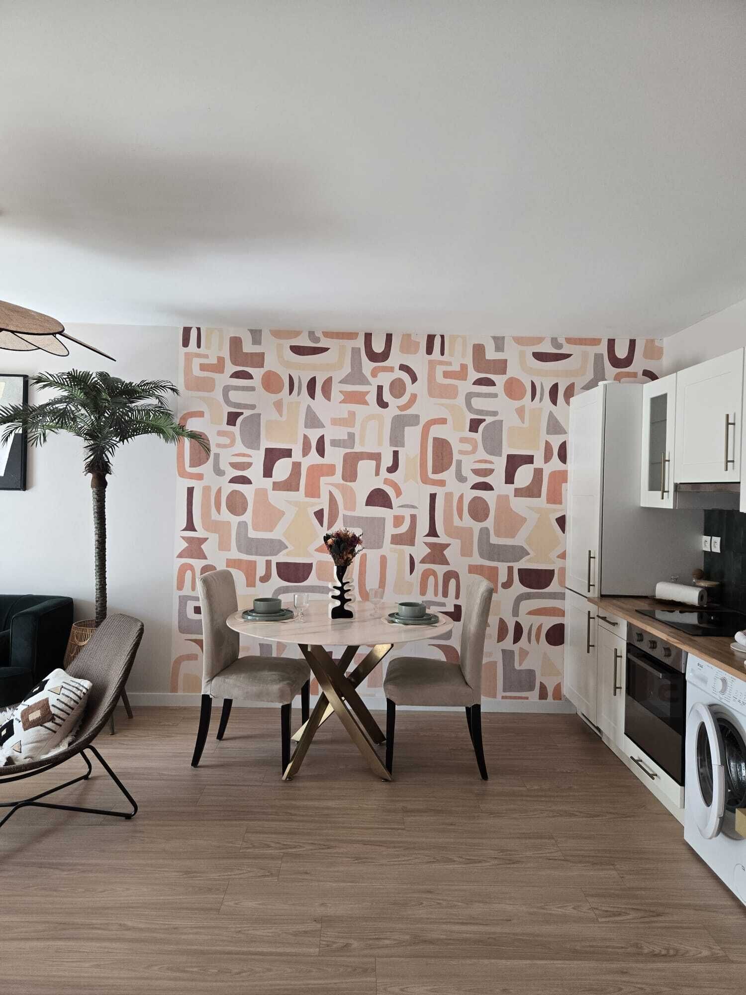 Eine moderne Küche mit weißen Schränken, einem Esstisch mit Stühlen, einer Waschmaschine und einer Wand mit abstrakten Mustern.