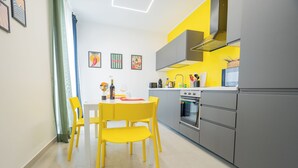 A modern kitchen with a dining area, featuring a white table, yellow chairs, and a grey refrigerator. - Colorama Home - Confort Loft  (Catania)