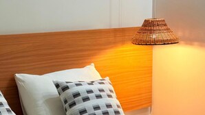 Una cama bien hecha con sábanas blancas y dos almohadas estampadas, cabecera de madera y lámpara de mimbre en la mesita de noche.