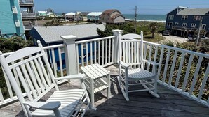 A wooden deck with white rocking chairs and a small table, overlooking a beach with buildings in the background.