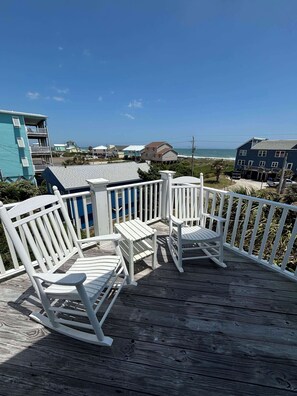 A wooden deck with white rocking chairs and a small table, overlooking a beach with buildings in the background. - NEW! Emerald Isle 2nd-Row Luxury 5BR/5+BA Home w/ Pool, Hot Tub & Ocean Views (Emerald Isle)