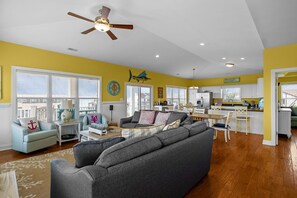 A spacious living room with a gray sectional sofa, a dining area, and a kitchen with white cabinets. - NEW! Emerald Isle 2nd-Row Luxury 5BR/5+BA Home w/ Pool, Hot Tub & Ocean Views (Emerald Isle)
