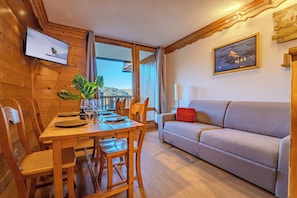 A wooden interior with a dining table, chairs, a sofa, a TV mounted on the wall, and a window with curtains. - La Roche Blanche Val Thorens (Les Belleville)