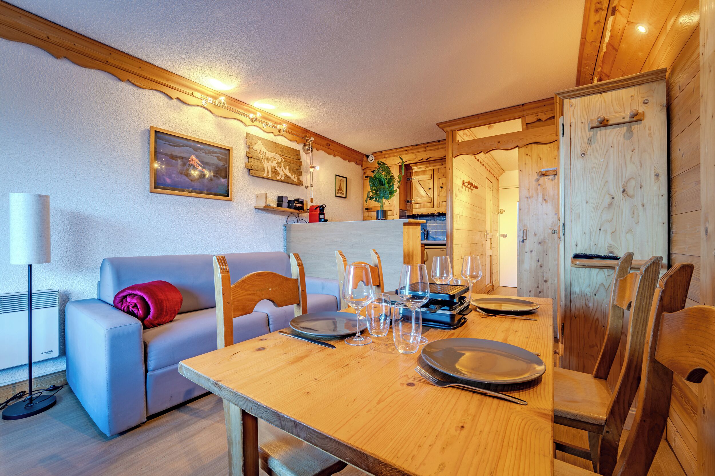 A cozy room with a wooden dining table set for four, a sofa with a red blanket, and a wall-mounted painting.