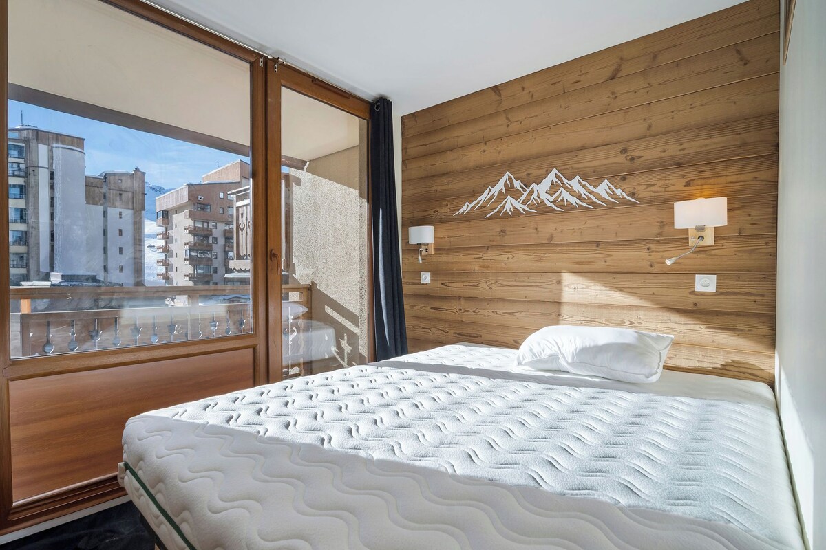 A hotel room with a bed, a wooden headboard, a window with a view of buildings, and a wall-mounted light fixture.