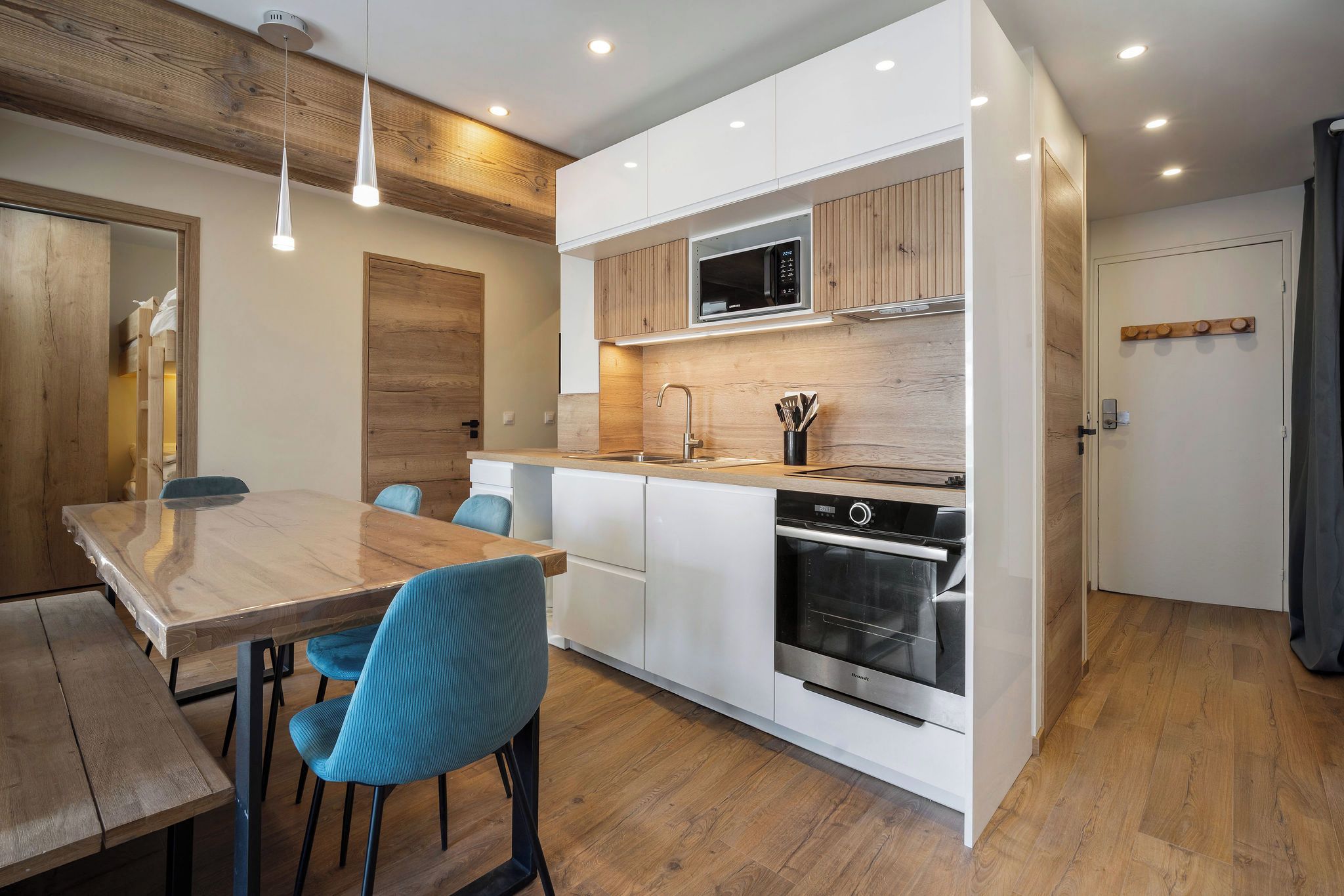 A modern kitchen with white cabinets, a built-in microwave, and a stainless steel oven. There is a wooden dining table with blue chairs.