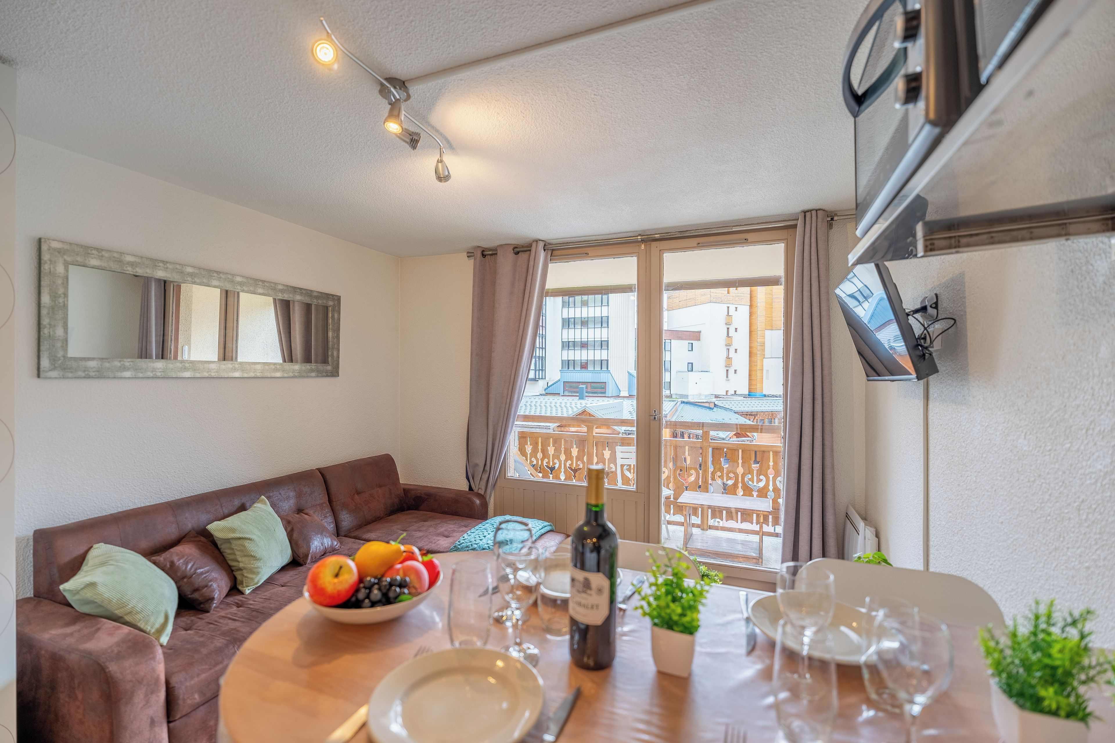Ein Wohnzimmer mit einem braunen Ledersofa, einem Esstisch gedeckt mit Tellern, Gläsern und einer Flasche, sowie einem großen Fenster mit Vorhängen.