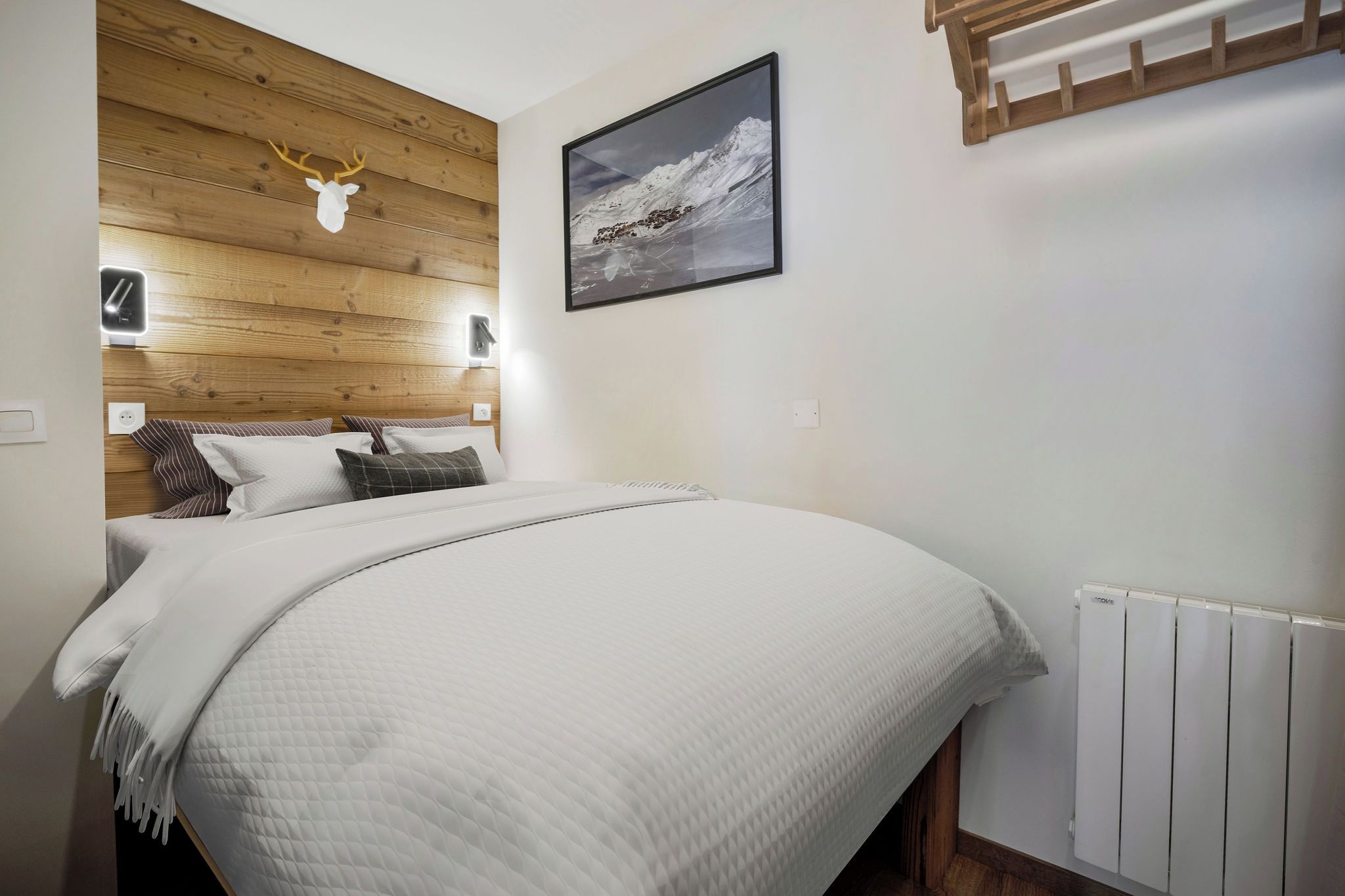 Quarto com cabeceira de madeira, cabeça de veado presa na parede, fotografia emoldurada de uma montanha, cama com roupa de cama branca e radiador branco.