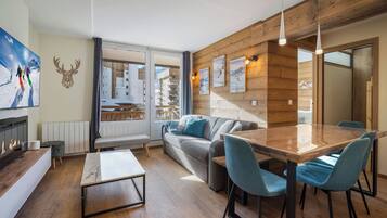 A modern living room with a wooden ceiling, a fireplace, a sofa, a dining table with chairs, and a view of a mountain through a glass door.