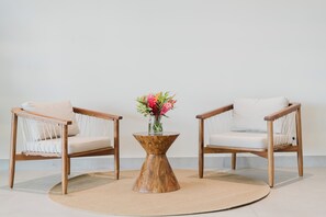 Dos sillas de madera con cojines, una mesa redonda de madera con un jarrón de flores, y un tapete beige sobre un suelo de baldosas.