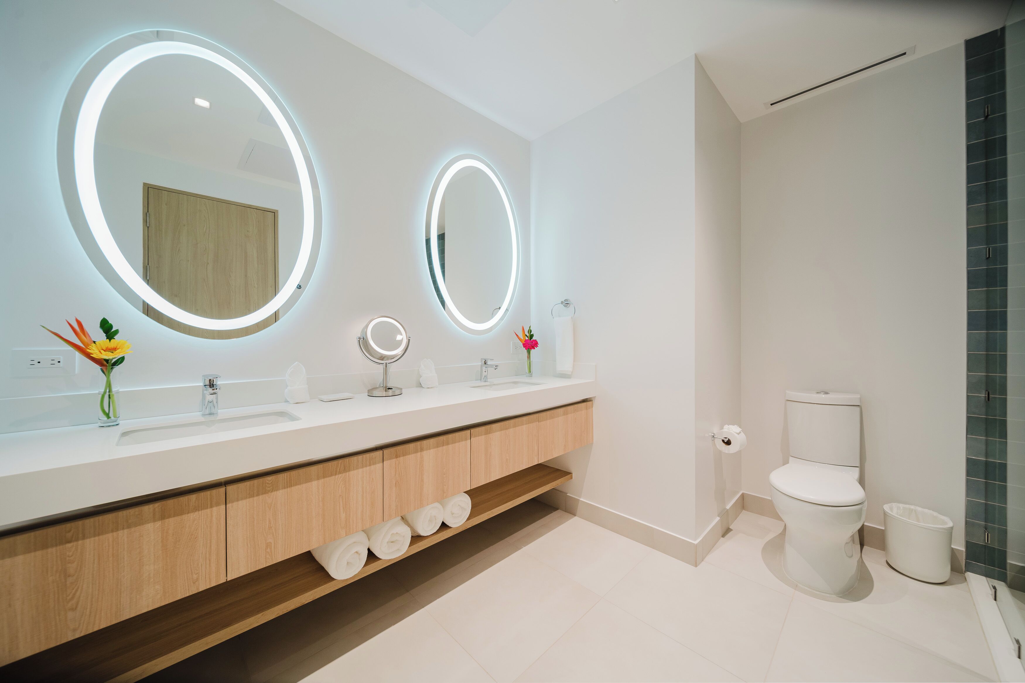 Un baño moderno con un tocador blanco, dos espejos iluminados, un inodoro y una zona de ducha.
