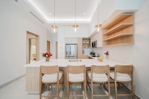 A modern kitchen with a central island, wooden chairs, pendant lights, and built-in shelving.