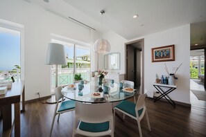 A modern dining area with a glass table, white chairs, a pendant light, and a view of the outdoors. - Discover the Pinnacle of Luxury and Serenity With La Costa! (Telire)