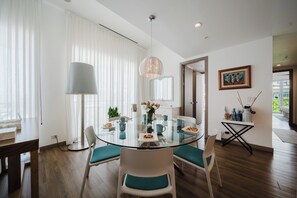 A modern dining area with a glass table, white chairs with teal cushions, a hanging light fixture, and a painting on the wall.