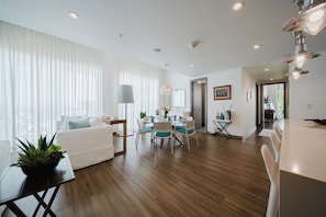 A modern living room with a dining area, wooden flooring, white furniture, and a large window with sheer curtains.
