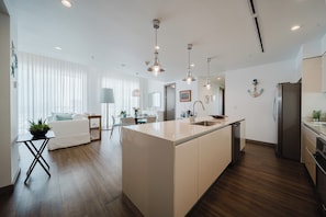 A modern kitchen with a central island, wooden flooring, pendant lighting, and a dining area with a white sofa and table.
