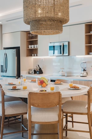 A modern kitchen with a round dining table, chairs, a fruit bowl, and a hanging light fixture. - Experience Expansive Luxury at our Gran Costa Villas! (Telire)