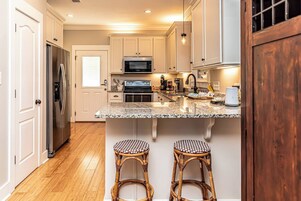 Una cocina con armarios blancos, electrodomésticos de acero inoxidable, encimera de granito y dos taburetes de mimbre.