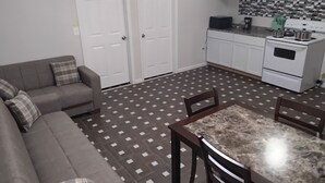 A living room with a sofa, a dining table, chairs, a kitchen with white cabinets, a stove, and a tiled floor. - Log Comfort 3 (Newark)