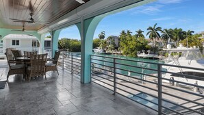 Ett takförsett utomhusområde med ett bord och stolar, en takfläkt och utsikt över en småbåtshamn med båtar och palmer.
