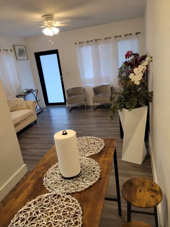 A modern living room with a wooden dining table, two chairs, a white vase with flowers, and a ceiling fan.